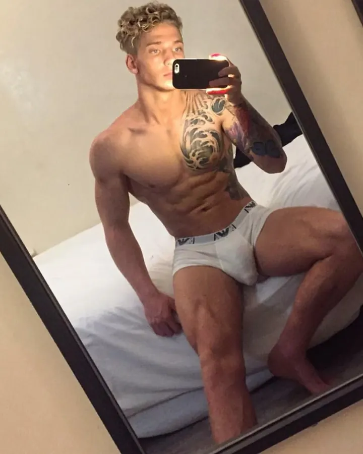 Homme torse nu, prenant un selfie dans un miroir, cheveux blonds bouclés, tatouages visibles sur le torse et le bras, en caleçon blanc.