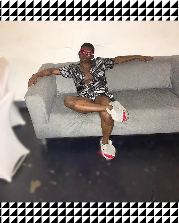 Homme en chemise rayée, portant des lunettes roses et des chaussons en forme de requin, assis sur un canapé gris.