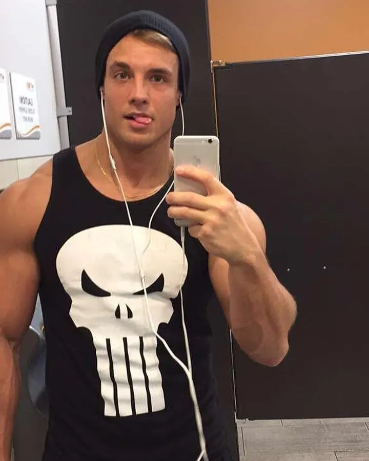Homme en t-shirt noir avec un logo de Punisher, prenant un selfie dans un gymnase, casquette noire, écouteurs blancs.