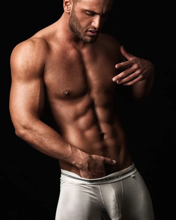 Homme torse nu, musclé, portant des sous-vêtements blancs, posant devant un fond noir.