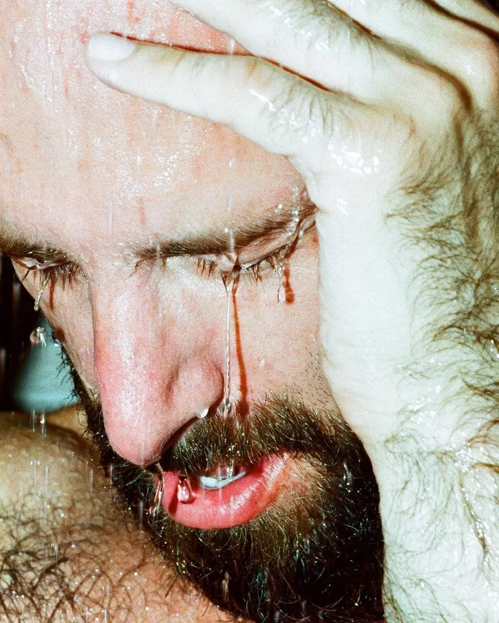 Plan rapproché d'un homme barbu, visage mouillé, main sur le front, eau coulant de ses yeux et de sa bouche.