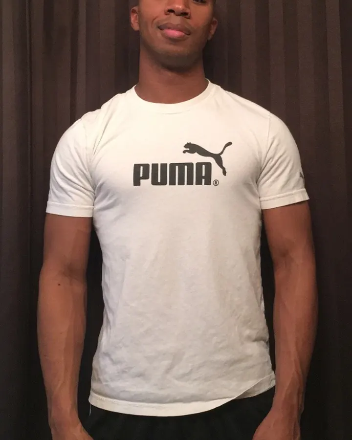Homme torse nu, t-shirt blanc Puma, sourire espiègle, debout devant un rideau sombre.