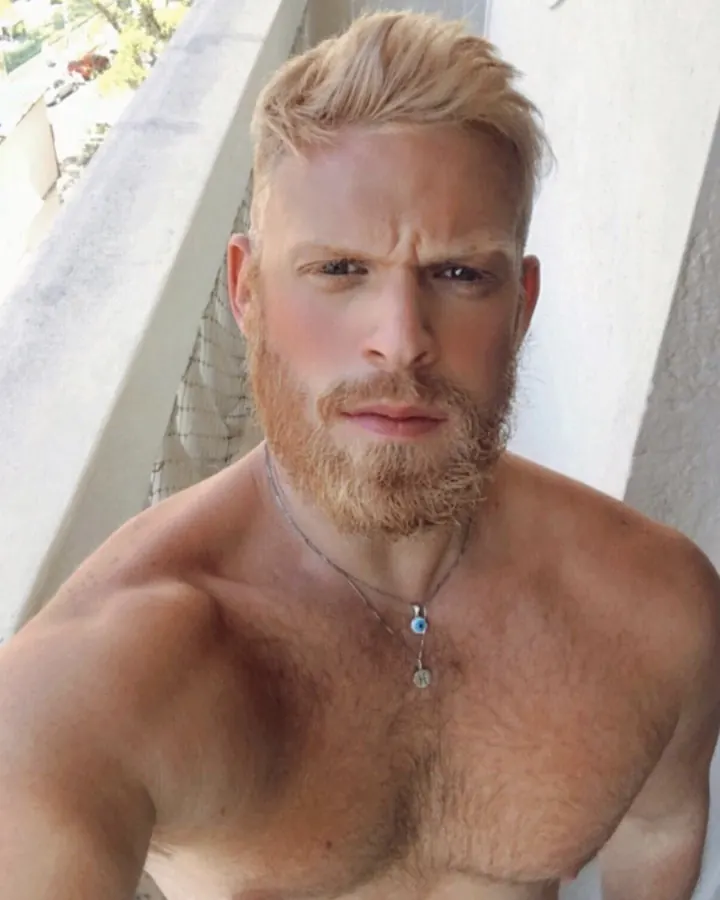 Homme barbu, cheveux blonds, torse nu, regard direct, en extérieur.
