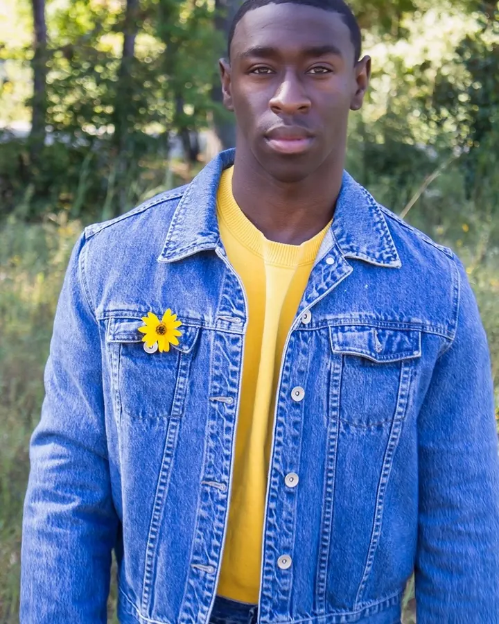 Homme en veste en jean bleu et t-shirt jaune, fleur jaune sur la poche, debout en extérieur.