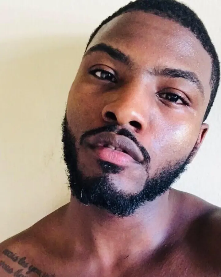 Homme torse nu, barbe courte, regard direct, tatouage visible sur la poitrine.