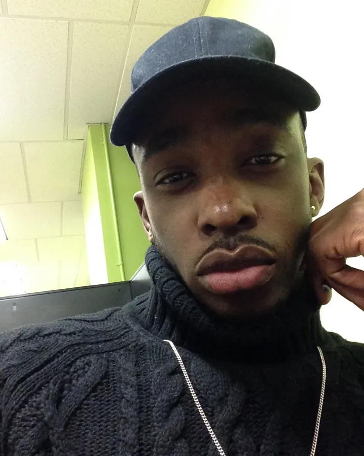 Homme en pull noir, casquette, col roulé, chaîne, regard direct, intérieur.