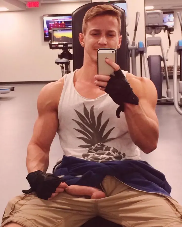 Homme musclé en t-shirt blanc avec motif ananas, prenant un selfie dans une salle de sport, portant des gants noirs