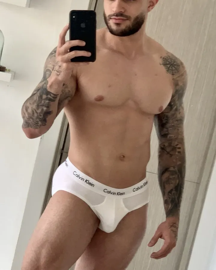 Homme torse nu en sous-vêtements blancs, tatouages aux bras, prend un selfie dans un miroir