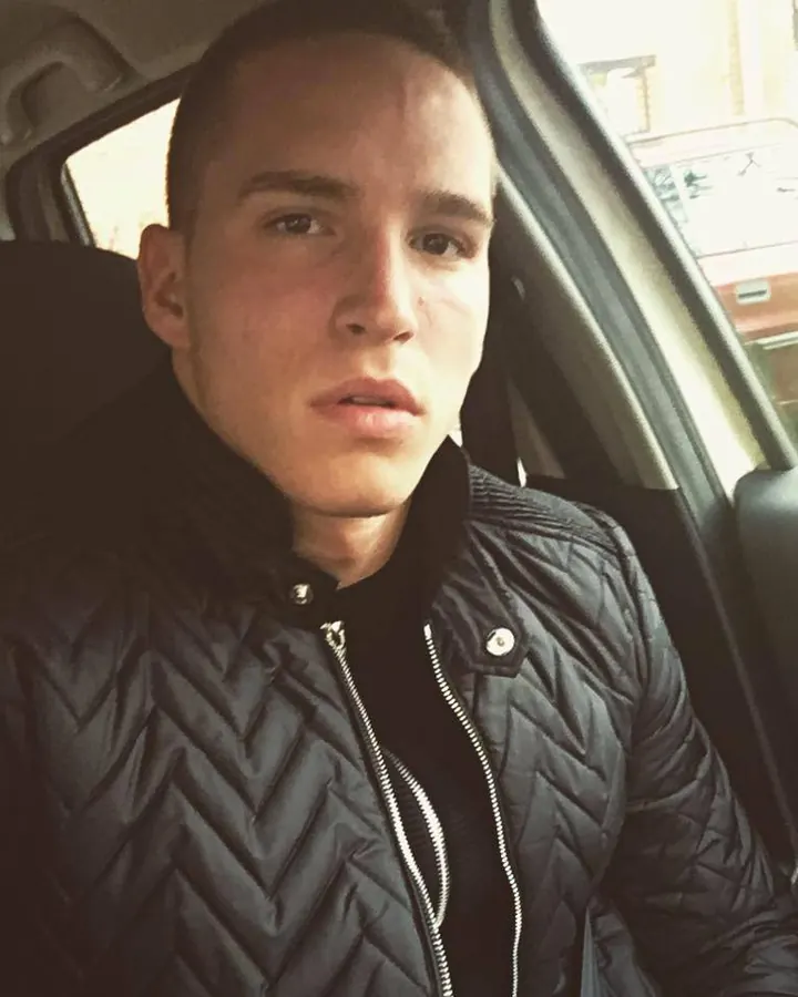Homme en veste noire, assis dans une voiture, regardant l'appareil photo avec une expression neutre.