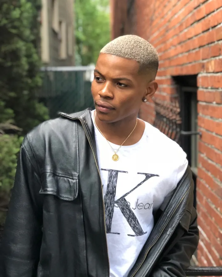 Homme africain avec cheveux platine, en veste en cuir noir, regard perçant contre un mur de briques