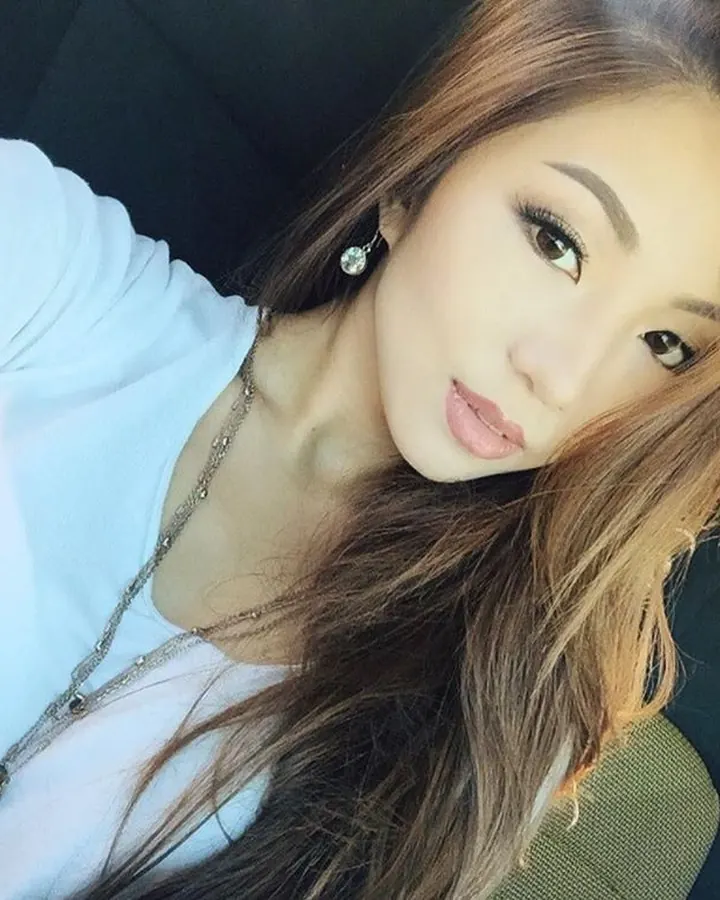 Jeune femme aux cheveux bruns, portant un haut blanc, assise dans une voiture, regardant l'appareil avec un sourire discret.